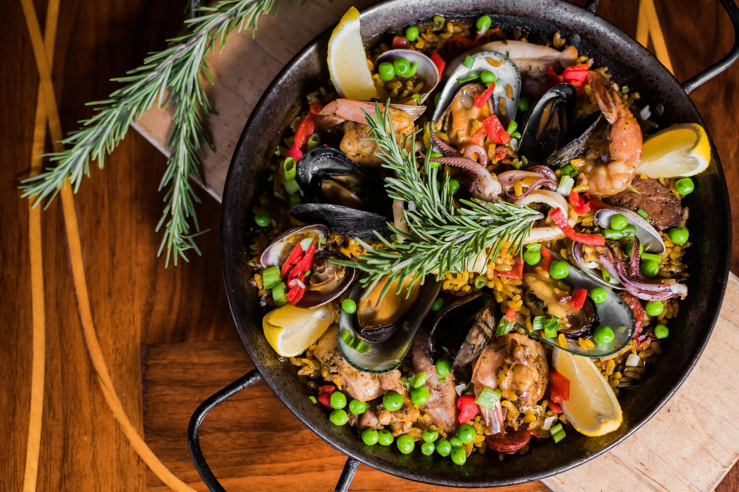 A pan of seafood paella with shrimp, mussels, clams, peas, red peppers, and lemon wedges, garnished with a sprig of rosemary, sits on a wooden table.