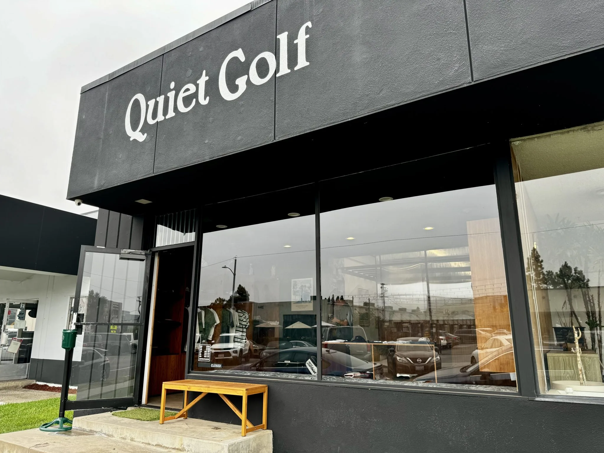 A storefront with the sign Quiet Golf above large windows, showing reflections of parked cars and buildings. An open door and a wooden bench are at the entrance. The sky appears overcast.