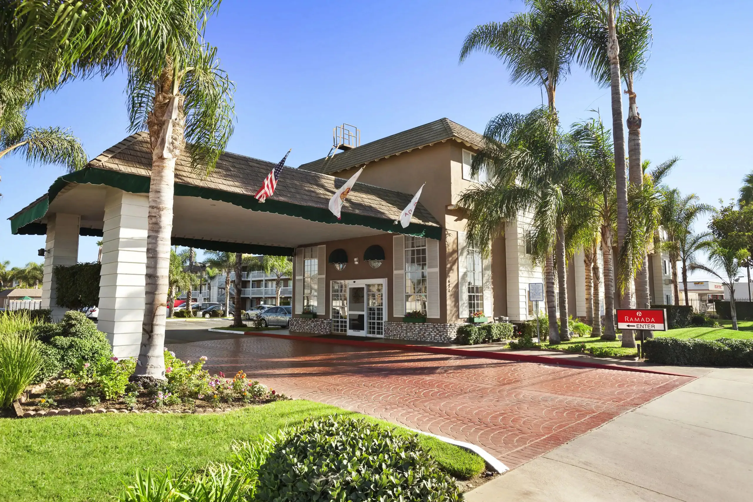 The Ramada Costa Mesa hotel entrance features a covered driveway, palm trees, three flags, pristine landscaping, and a clear blue sky. The light-colored building offers large windows and a red brick-patterned drive for a welcoming arrival.