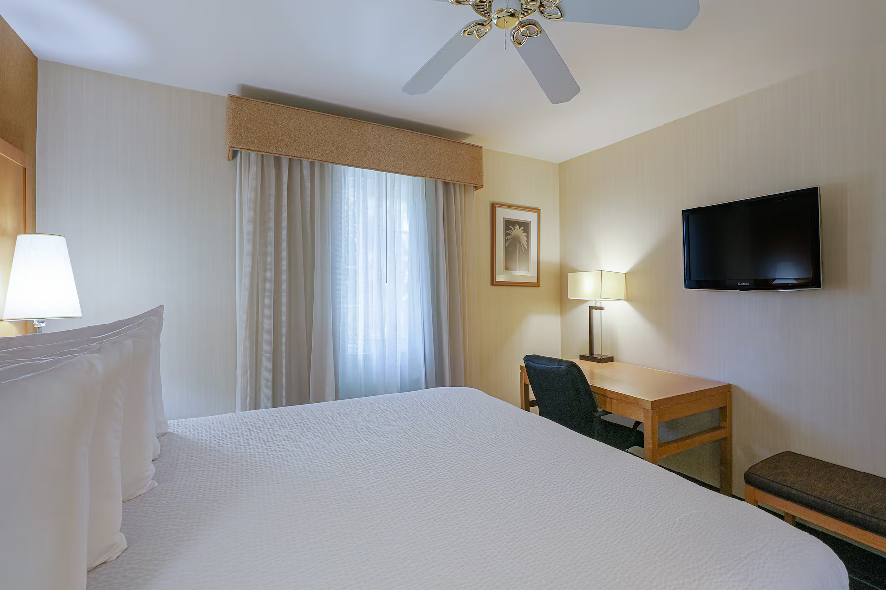 Hotel room at Ramada Costa Mesa with a neatly made bed, wall-mounted TV, desk and chair, a window with curtains, ceiling fan, lamps, and framed artwork. The space is clean and softly lit for your comfort.