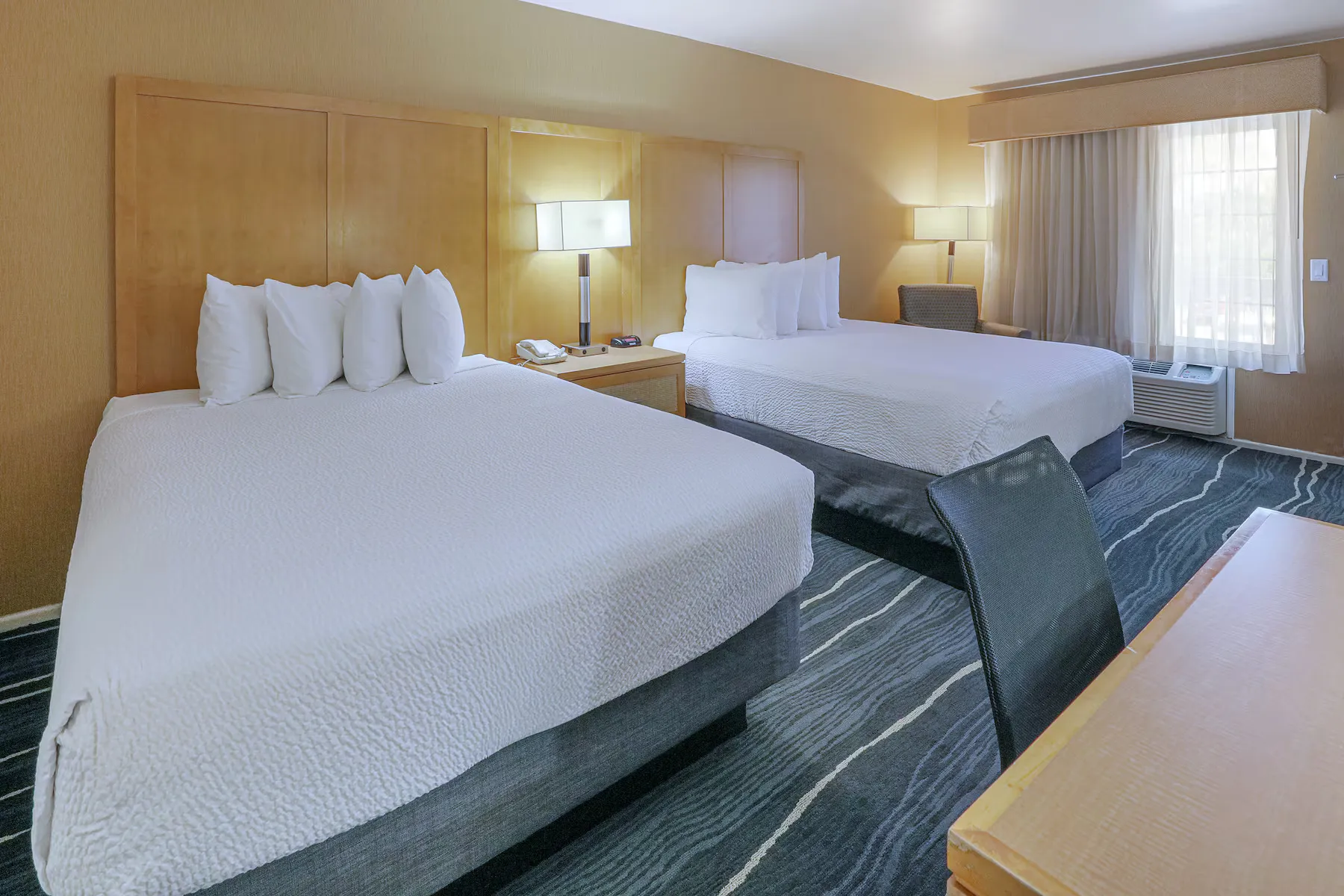 A Ramada Costa Mesa hotel room with two large beds, crisp white bedding, a bedside table with lamp and phone, a work desk with chair, and a window with curtains letting in natural light.