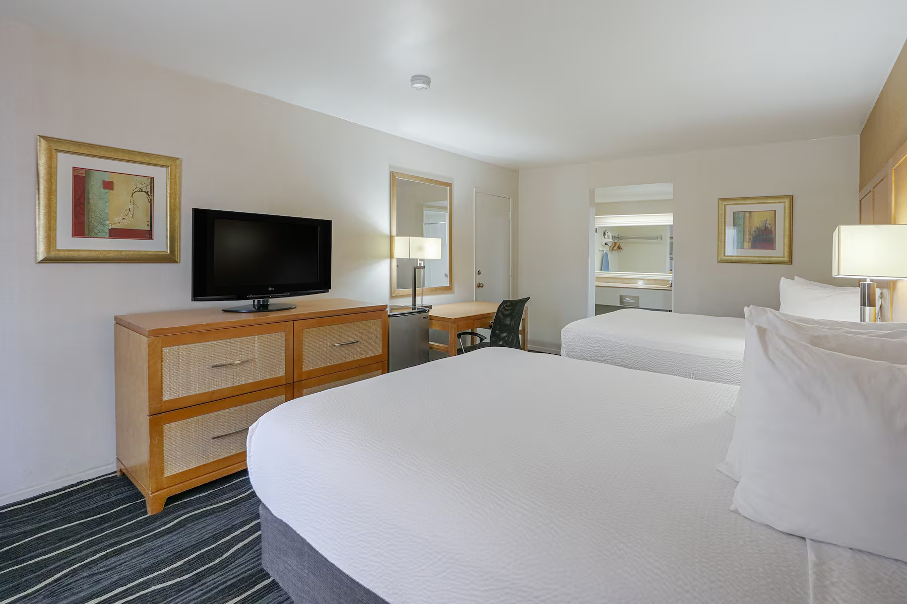 A Ramada Costa Mesa hotel room with two double beds, white bedding, a TV on a wooden dresser, a desk with a chair, lamp, framed artwork on the walls, and an open vanity area with a mirror in the background.