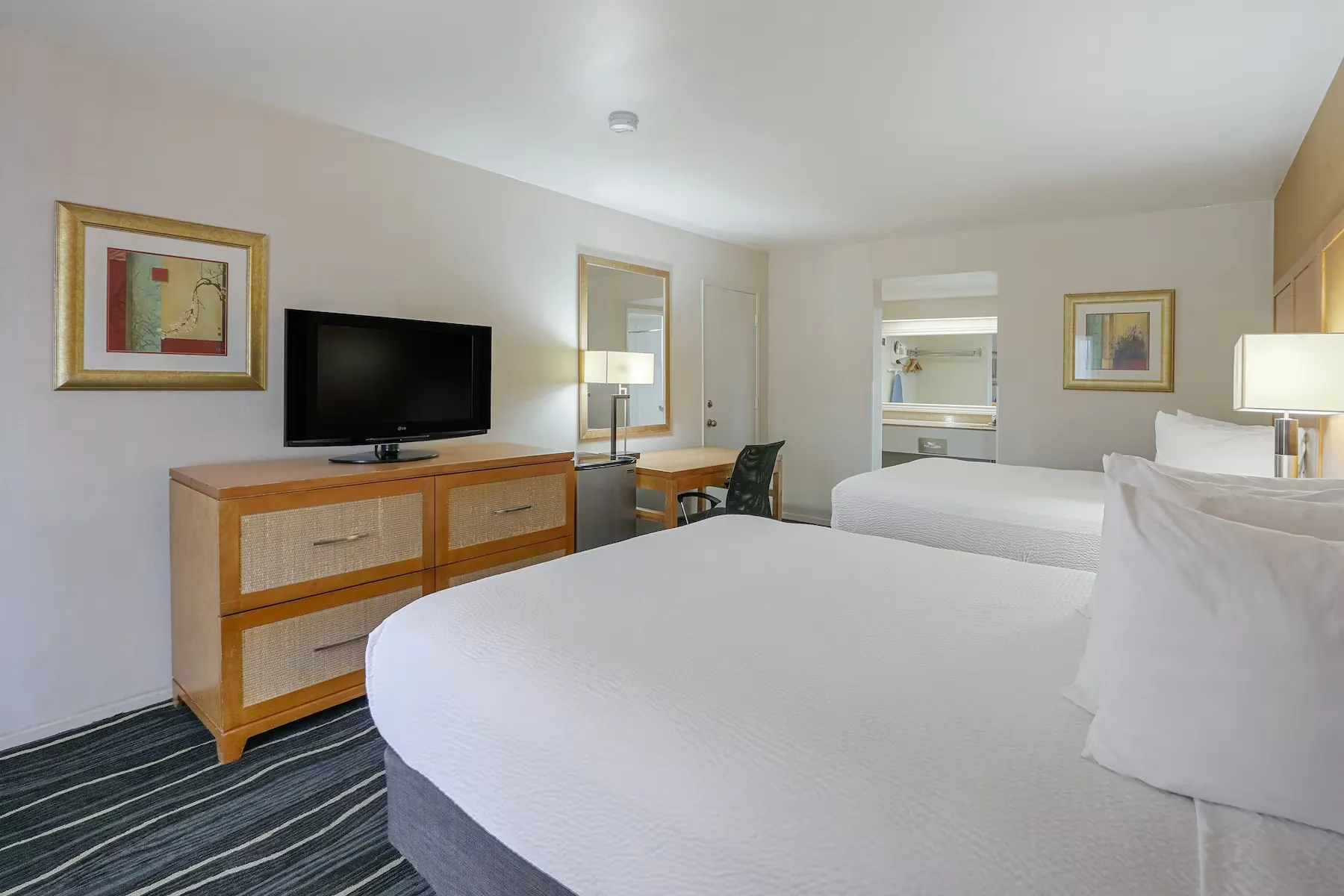 A Ramada Costa Mesa hotel room with two double beds, white bedding, a TV on a wooden dresser, a desk with a chair, lamp, framed artwork on the walls, and an open vanity area with a mirror in the background.