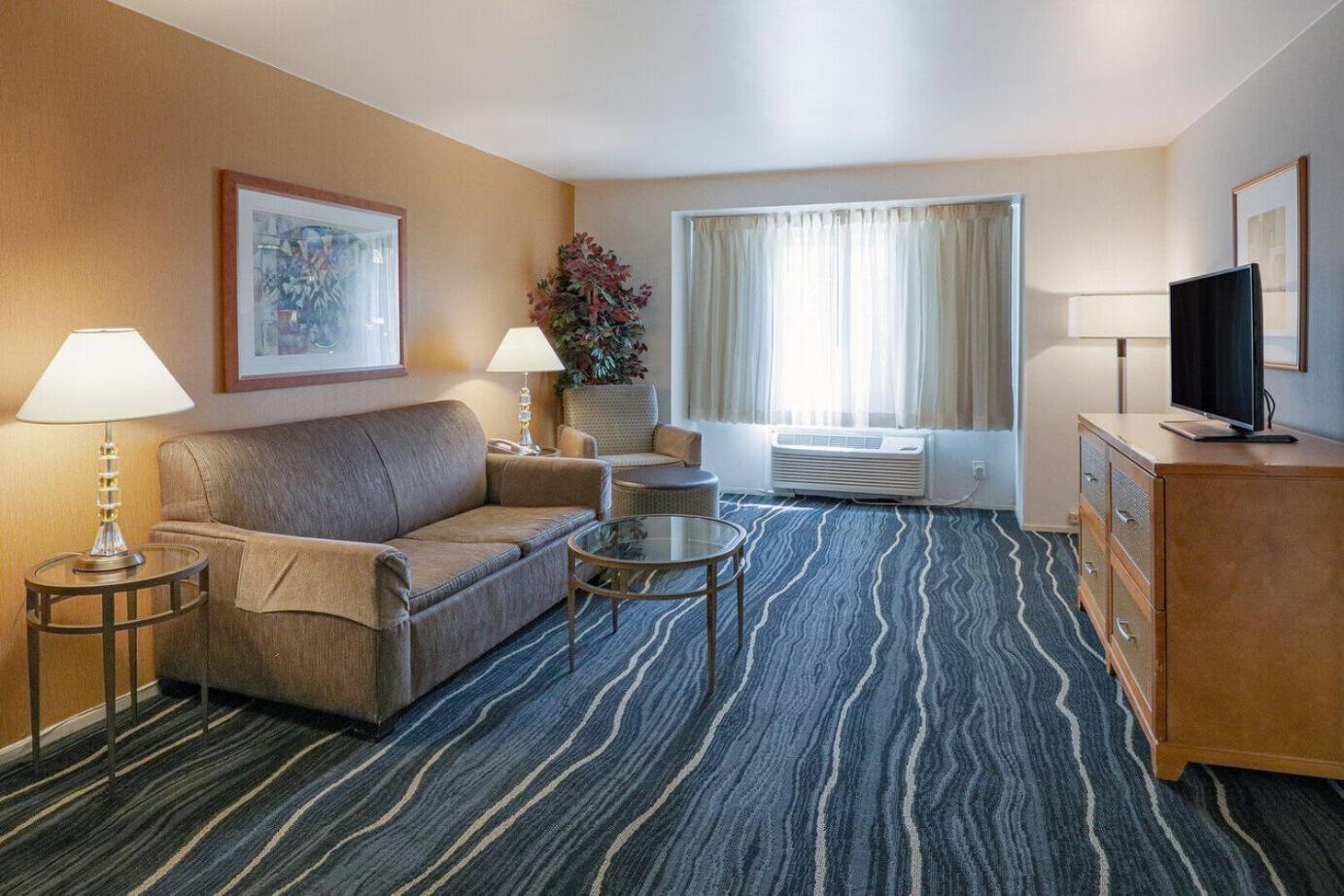 A hotel room at Ramada Costa Mesa features a brown sofa, glass coffee table, armchair, TV on a wooden dresser, and two lamps. The space boasts a patterned carpet, large window with curtains, wall art, and a potted plant in the corner.