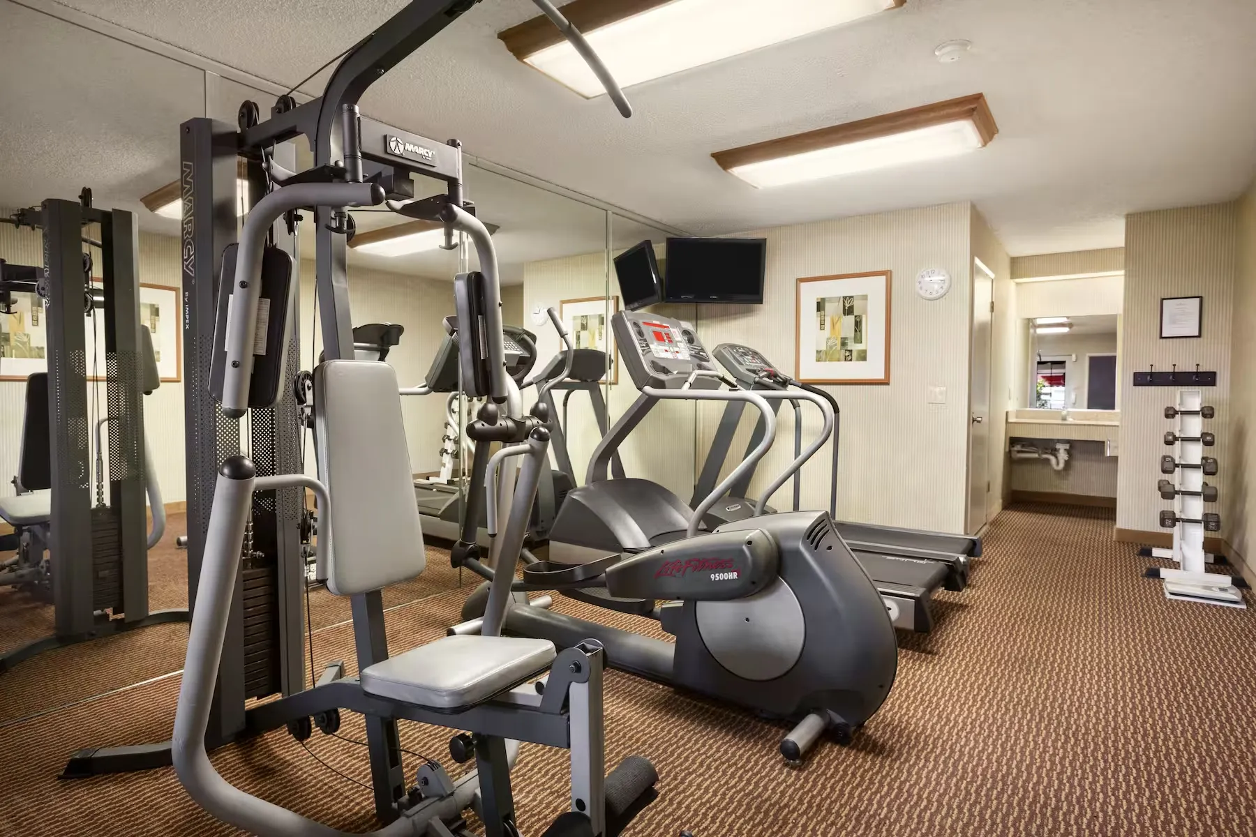 A small gym room at Ramada Costa Mesa with a weight machine, two treadmills, free weights, wall mirrors, framed art, a wall clock, and a TV mounted above the equipment.