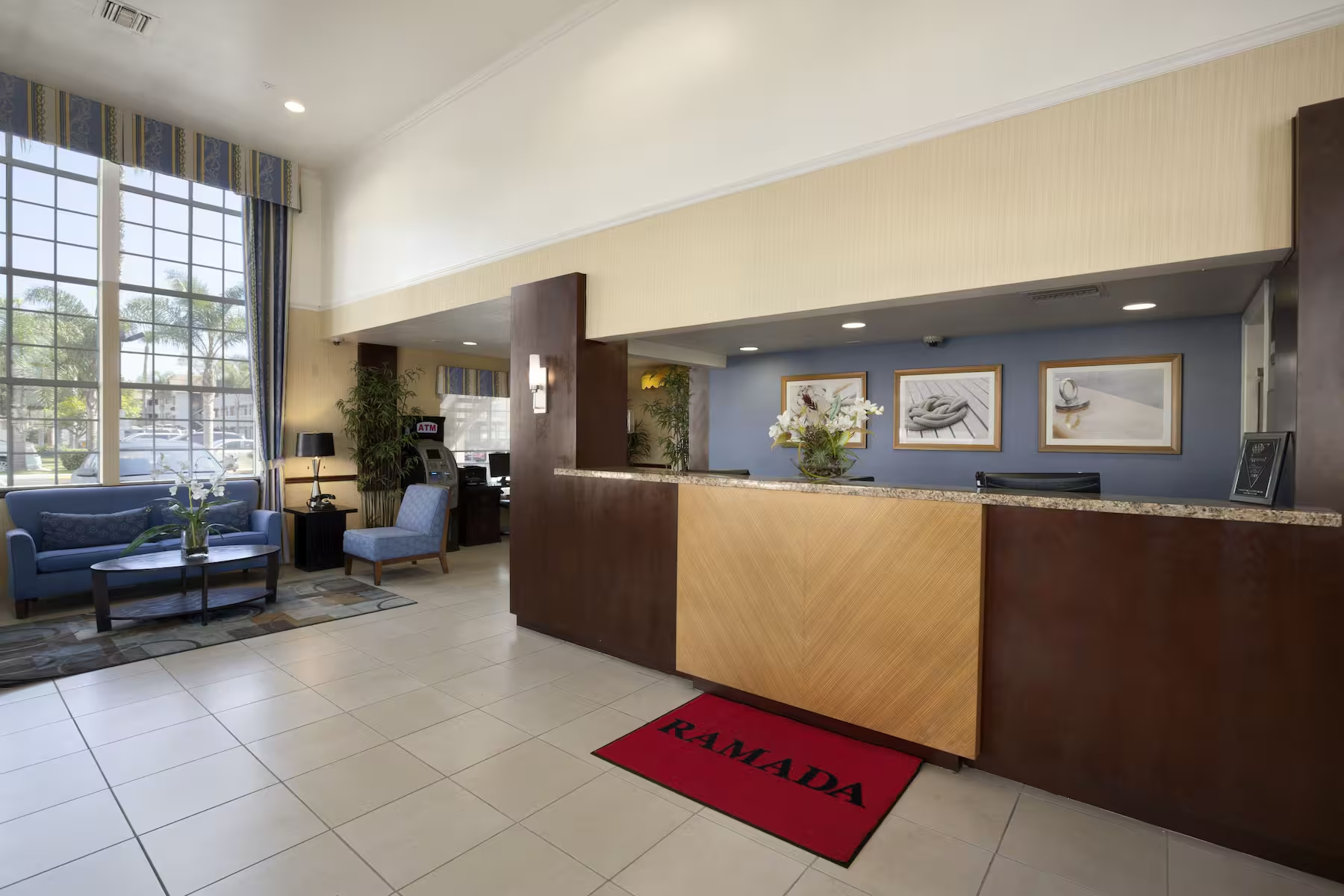 A hotel lobby at Ramada Costa Mesa features a reception desk, a red Ramada mat, blue couches, large windows filling the space with natural light, and framed artwork on the wall behind the counter.