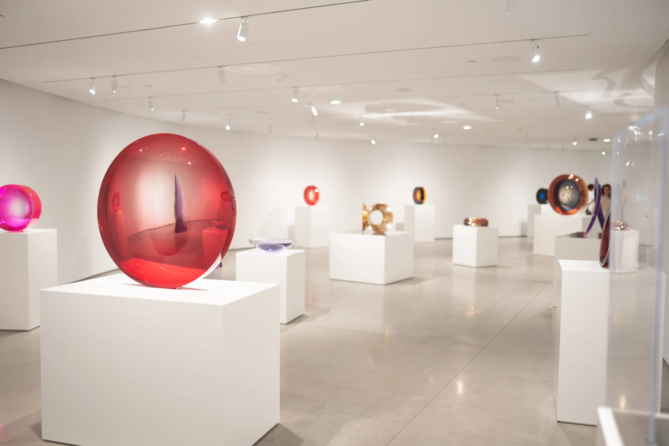 Modern art gallery with large, colorful glass sculptures displayed on white pedestals in a bright, spacious room with white walls and polished concrete floors, reminiscent of the contemporary exhibitions at the Orange County Museum of Art.