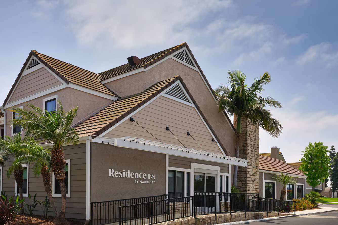 A beige and tan building with a peaked roof, palm trees, and “Residence Inn by Marriott” signage on the front welcomes you to Residence Inn Costa Mesa, set beneath a blue sky with scattered clouds.