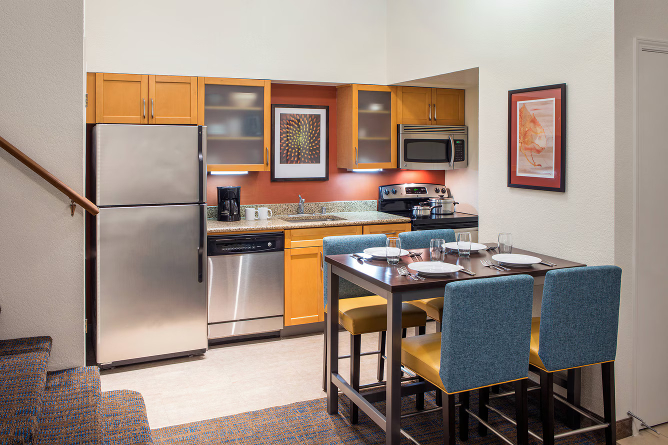 Modern kitchen at Residence Inn Costa Mesa with wooden cabinets, stainless steel appliances, a dining table set for four with blue and yellow chairs, and a staircase on the left. A framed artwork hangs on the white wall.