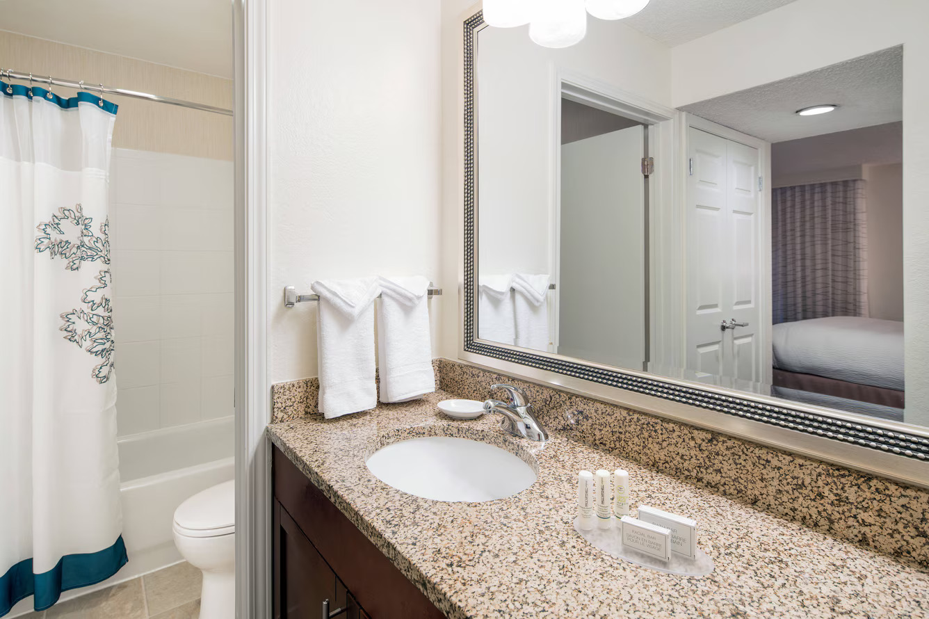 A clean bathroom at Residence Inn Costa Mesa features a granite countertop, sink, mirror, white towels on a rack, and toiletries. In the background, there's a bathtub with a shower curtain and a glimpse of the bedroom through an open door.
