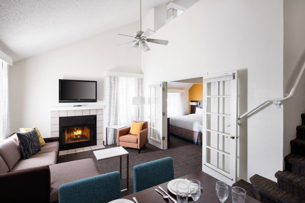 A modern hotel suite at Residence Inn Costa Mesa features a living area with a sofa, armchair, fireplace, and TV. The dining table is set for guests. French doors open to the bedroom, and a staircase adds to the spacious layout.