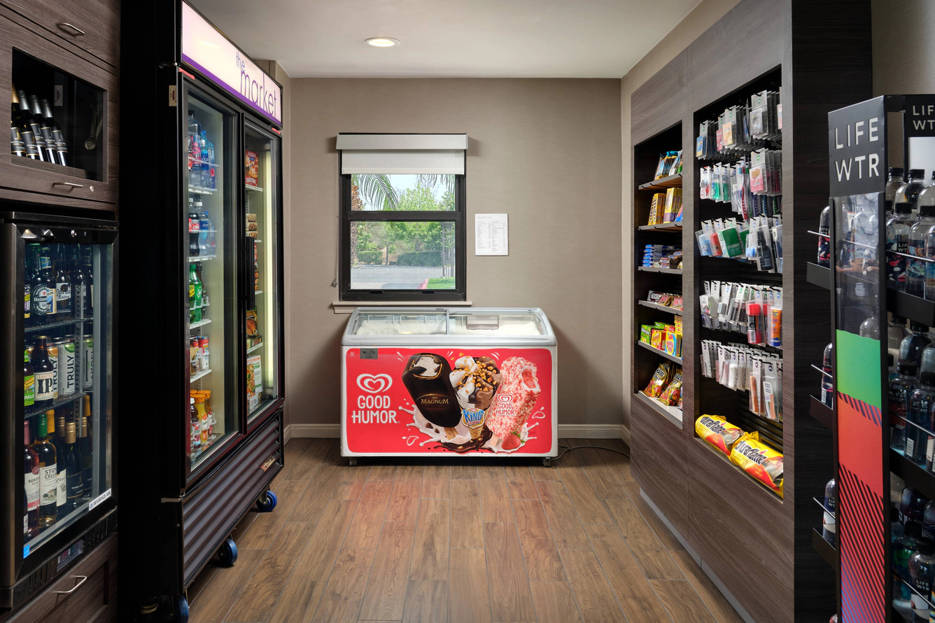 A small convenience store at Residence Inn Costa Mesa with shelves of snacks, a fridge stocked with drinks, a Good Humor-branded ice cream freezer, and a window in the back letting in natural light.