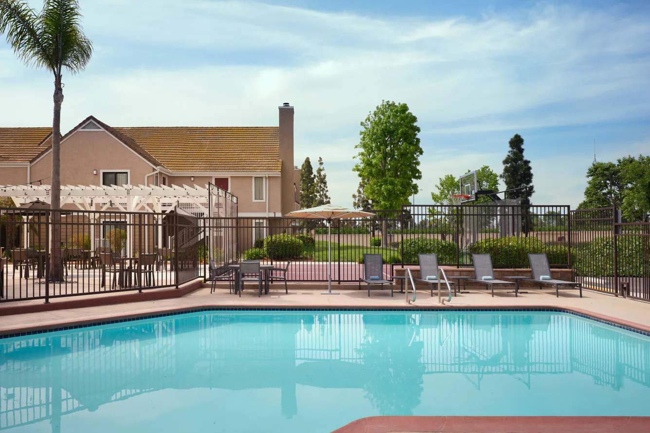 A clear outdoor swimming pool at the Residence Inn Costa Mesa is surrounded by lounge chairs and tables, fenced in with a basketball hoop and green trees in the background, next to a tan building under a partly cloudy sky.