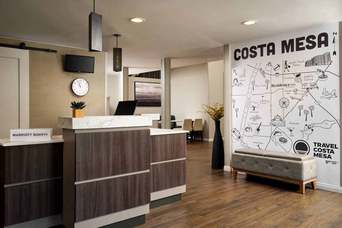 Modern hotel lobby with a front desk, a clock on the wall, a Marriott Bonvoy sign, and a large illustrated map of Costa Mesa next to a gray bench at the Residence Inn Costa Mesa.
