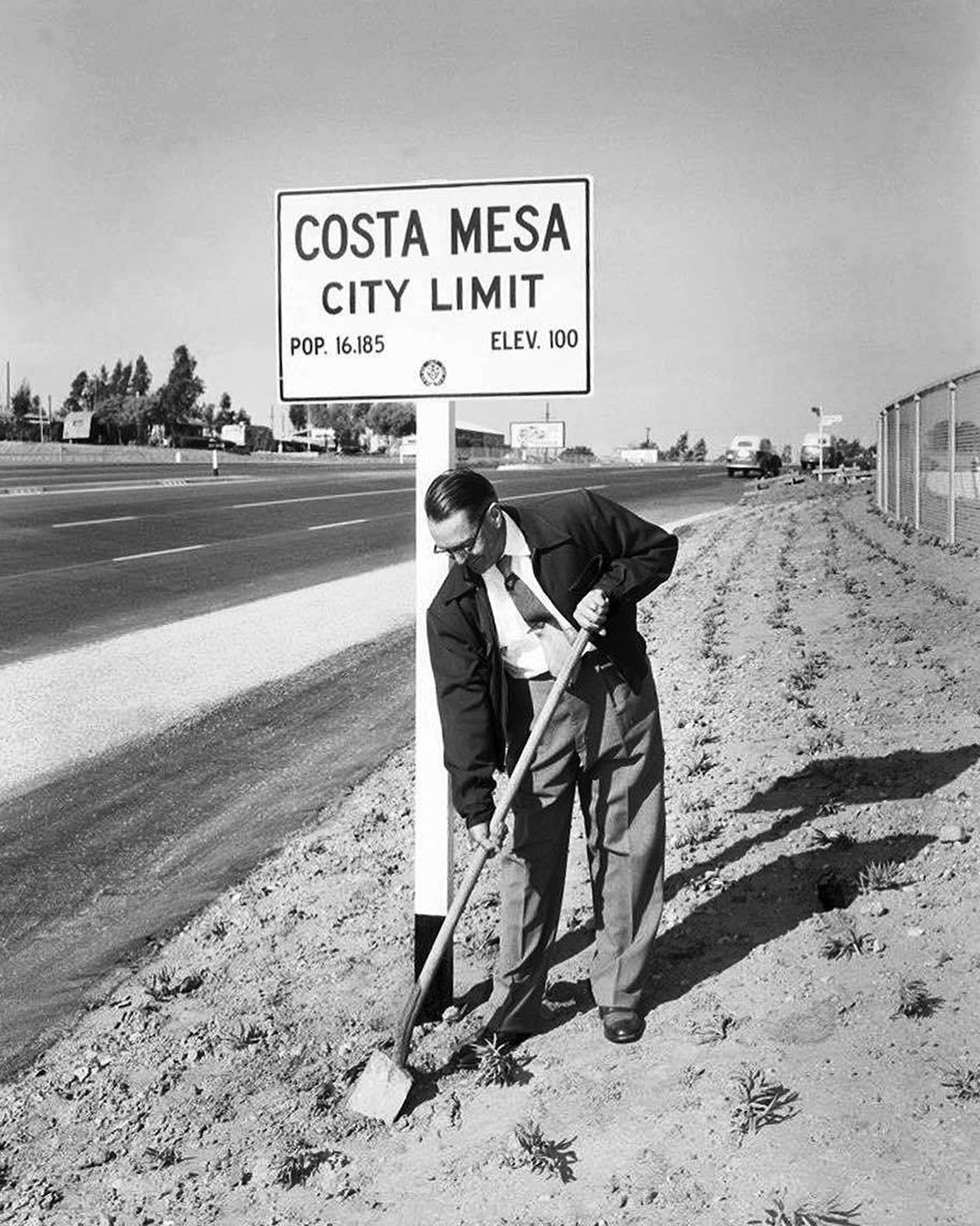 A man in a suit and jacket uses a shovel to dig near a road by the COSTA MESA CITY LIMIT sign. This vintage scene captures the spirit of mid-20th century Costa Mesa—perfect inspiration for a costa mesa travel blog.