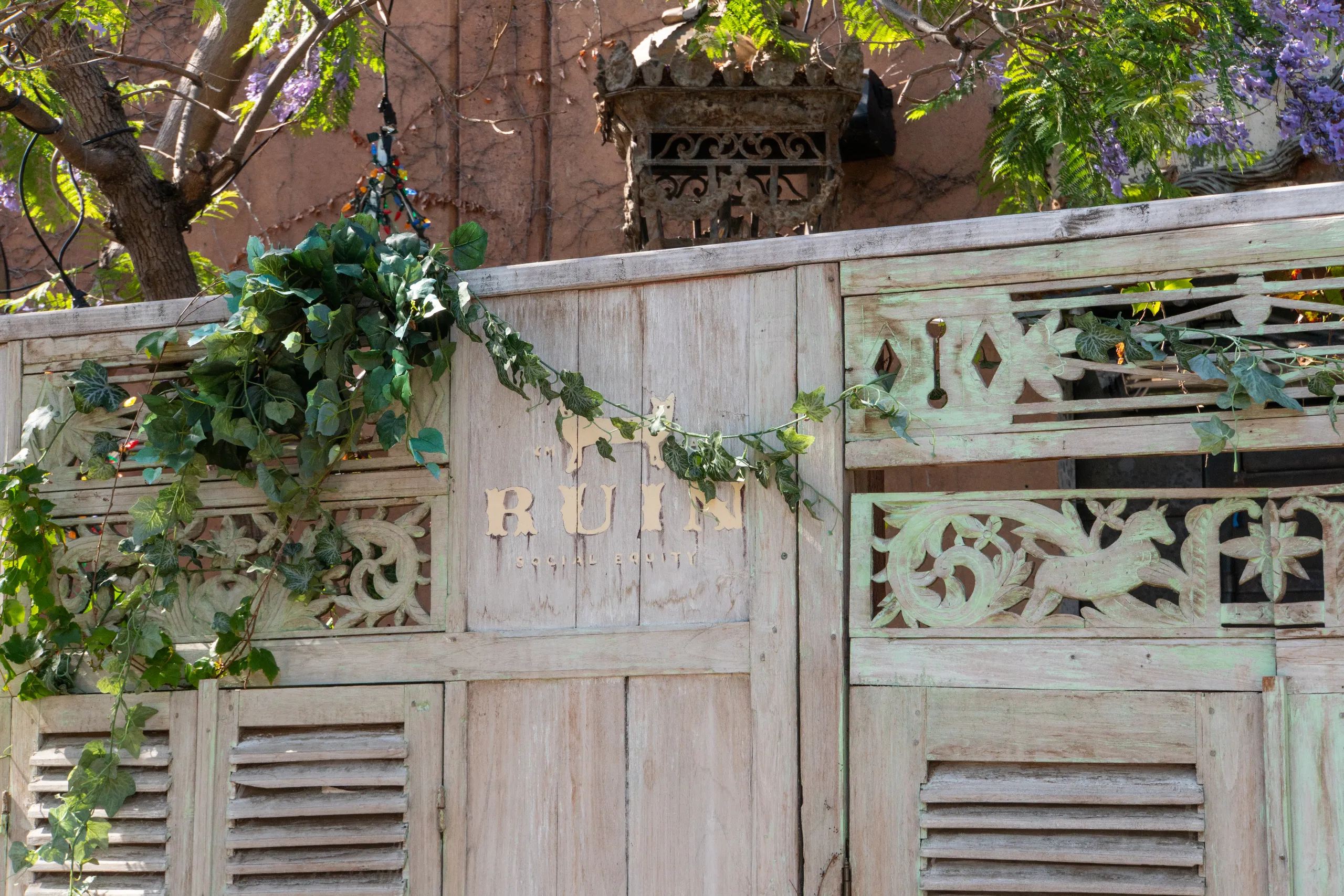 A rustic wooden gate with intricate carvings and greenery draped over it. The faded letters RUIN are visible on the gate. Trees with green leaves and purple flowers are in the background.