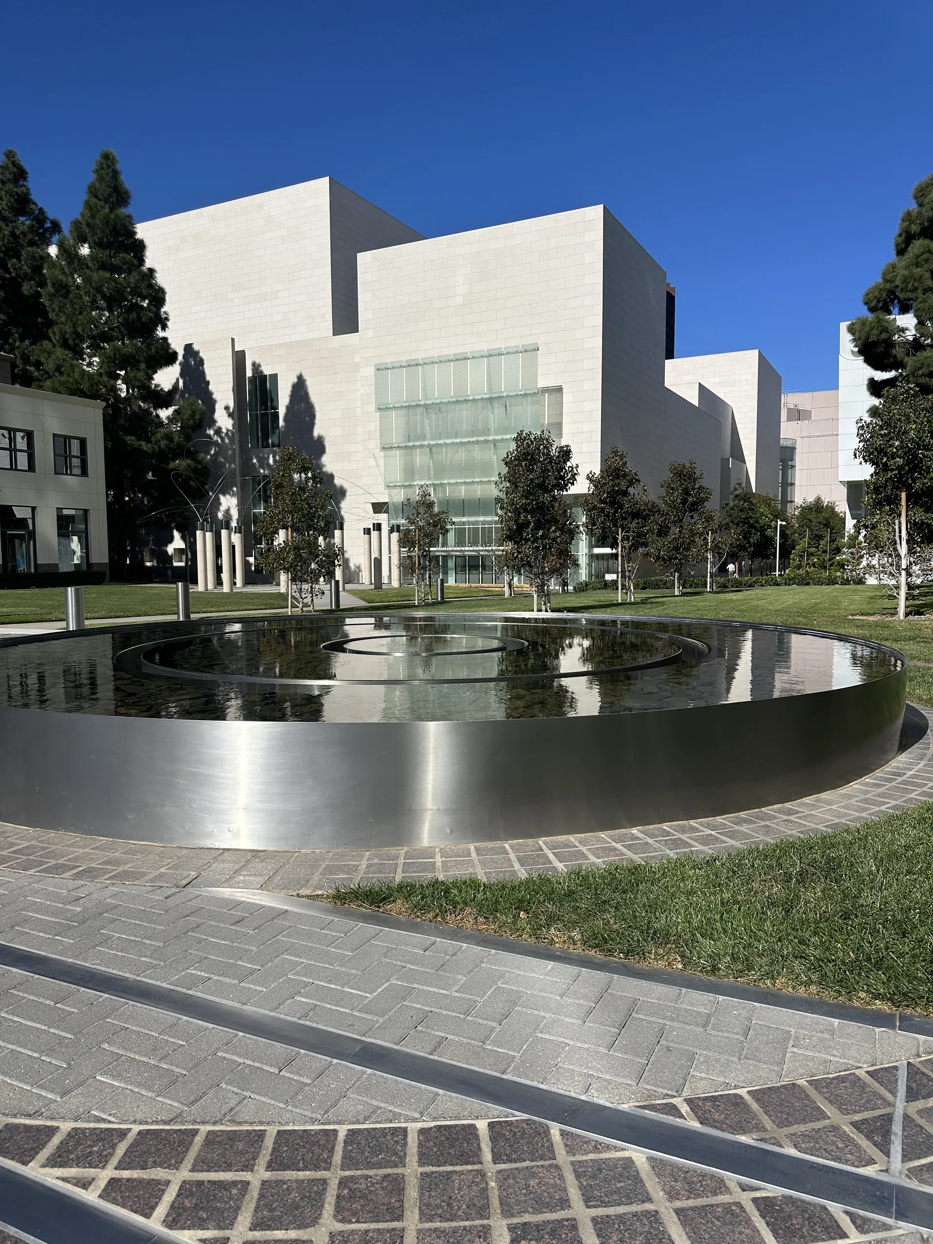 A modern circular reflecting pool with a metallic edge sits in front of light-colored contemporary buildings and trees near the Samueli Theater in Costa Mesa, under a clear blue sky.