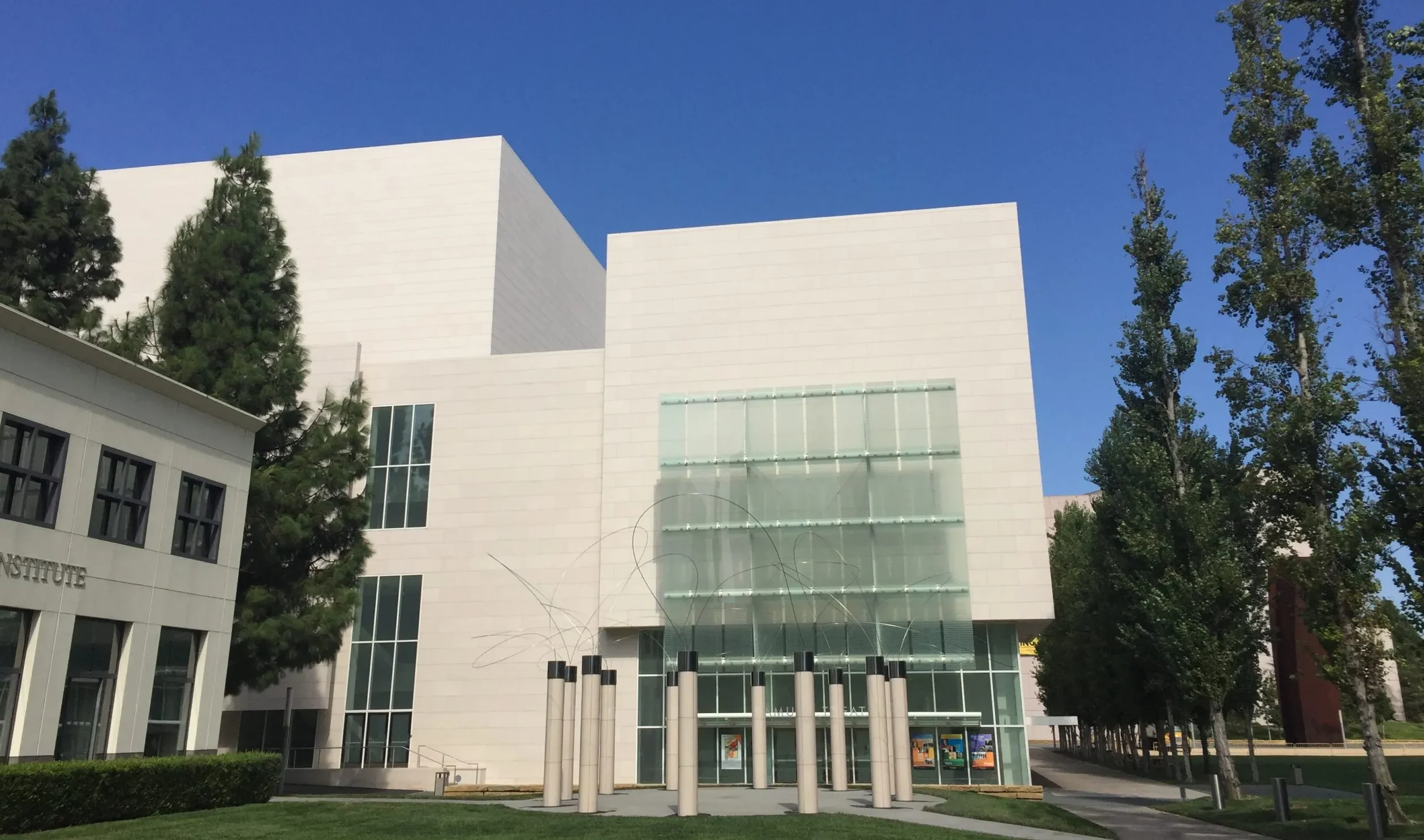Modern white building with glass entrance, metal sculpture in front, green lawn, trees, and a clear blue sky—just steps from Samueli Theater Costa Mesa.