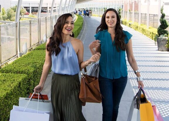Two women smiling and walking outside on a sunny day, each carrying several shopping bags on a pedestrian bridge lined with green bushes and metal railings.