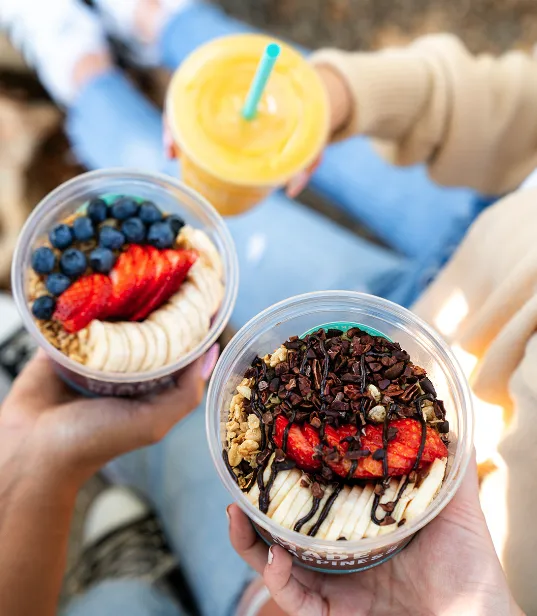 Two people holding smoothie bowls topped with banana slices, strawberries, granola, and blueberries or chocolate drizzle; one person also holds a yellow smoothie with a blue straw. They are sitting outdoors in casual clothing.