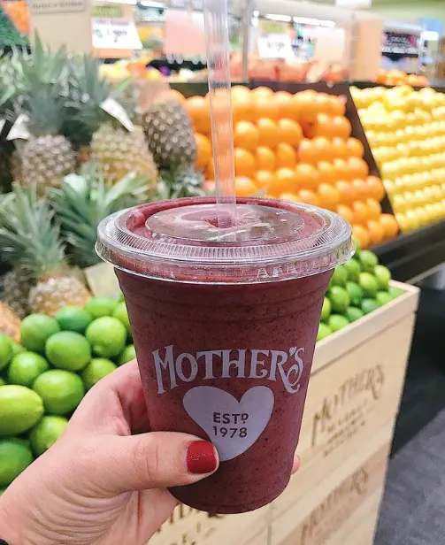 A hand with red nail polish holds a smoothie in a clear cup labeled Mother’s in front of a grocery display of pineapples, limes, and oranges.