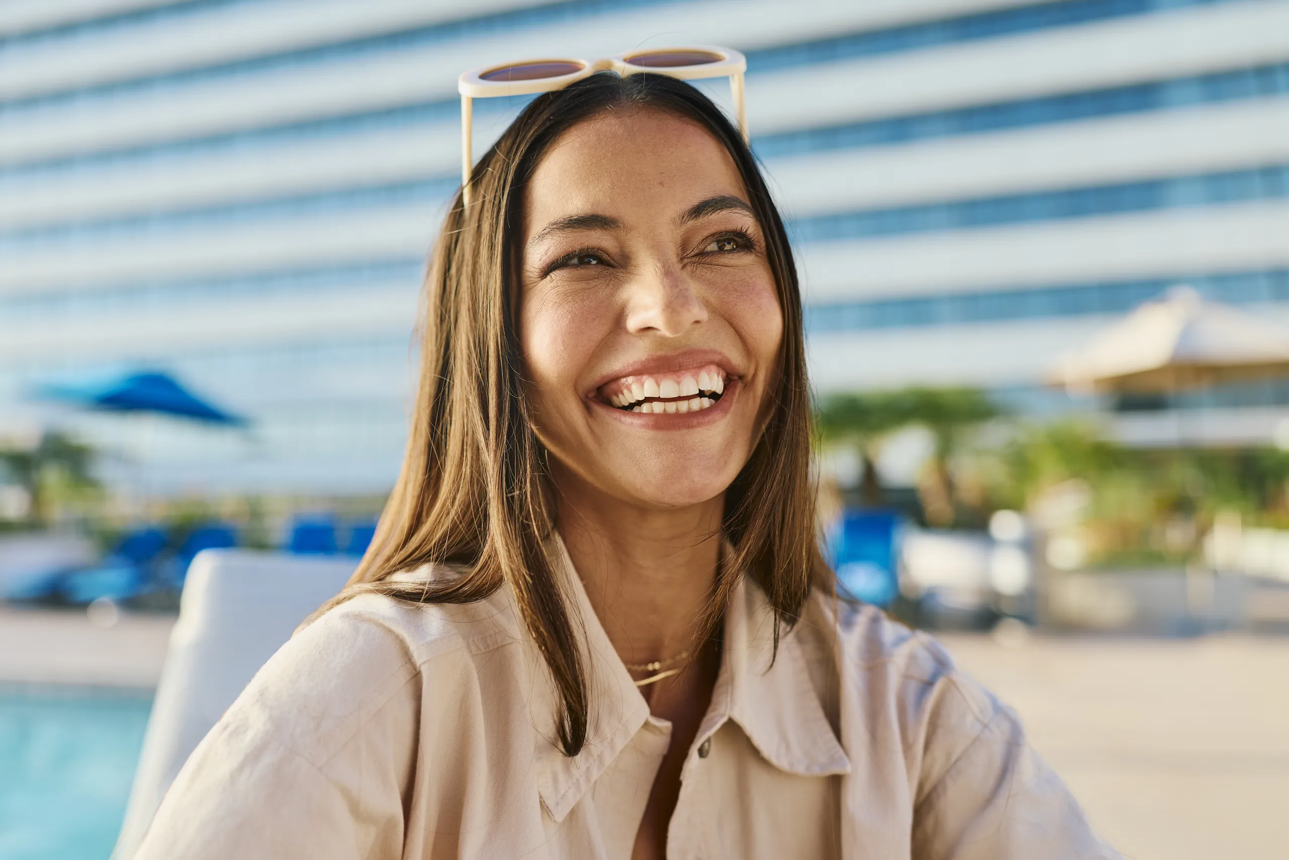 A smiling woman with long brown hair, wearing a light-colored button-up shirt and sunglasses resting on her head, looks to the side with a bright, joyful expression—perfect for a Costa Mesa travel blog. The background shows a blurry swimming pool area and a modern building.