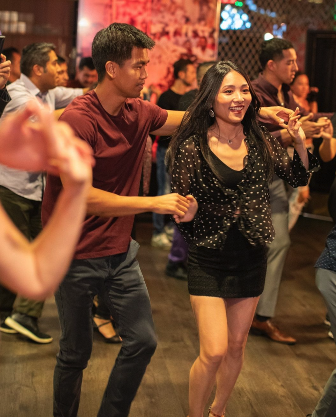 A man and a woman dance together in a lively, crowded indoor setting. The woman smiles, wearing a black outfit, while the man leads her. Other people dance in the background, creating an energetic atmosphere.