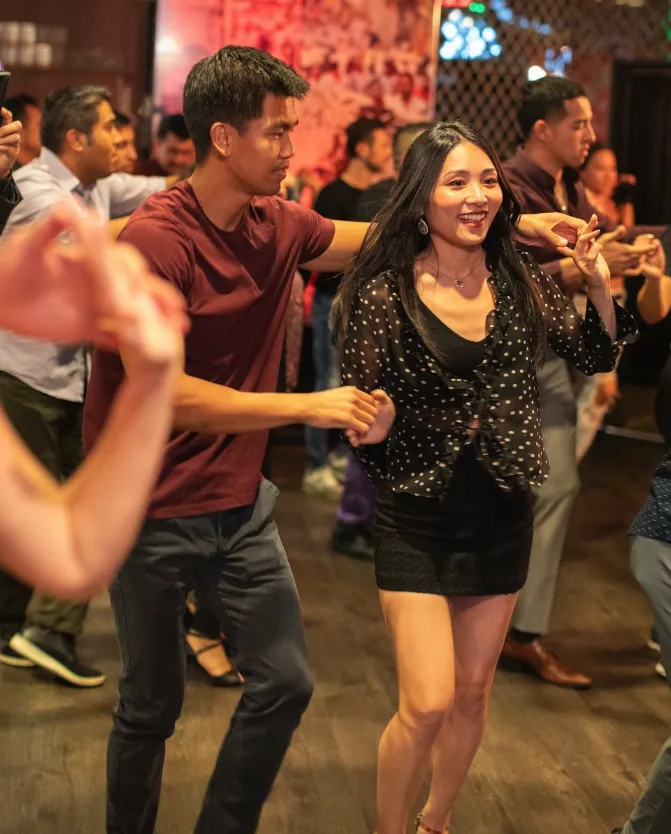 A man and a woman dance together in a lively, crowded indoor setting. The woman smiles, wearing a black outfit, while the man leads her. Other people dance in the background, creating an energetic atmosphere.