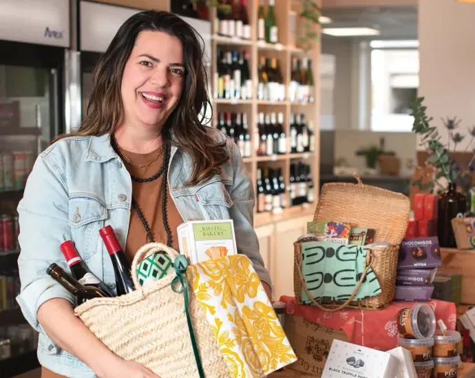 A smiling woman stands in a store holding a gift basket filled with wine and other items. Shelves of wine bottles and various gift baskets are visible in the background.