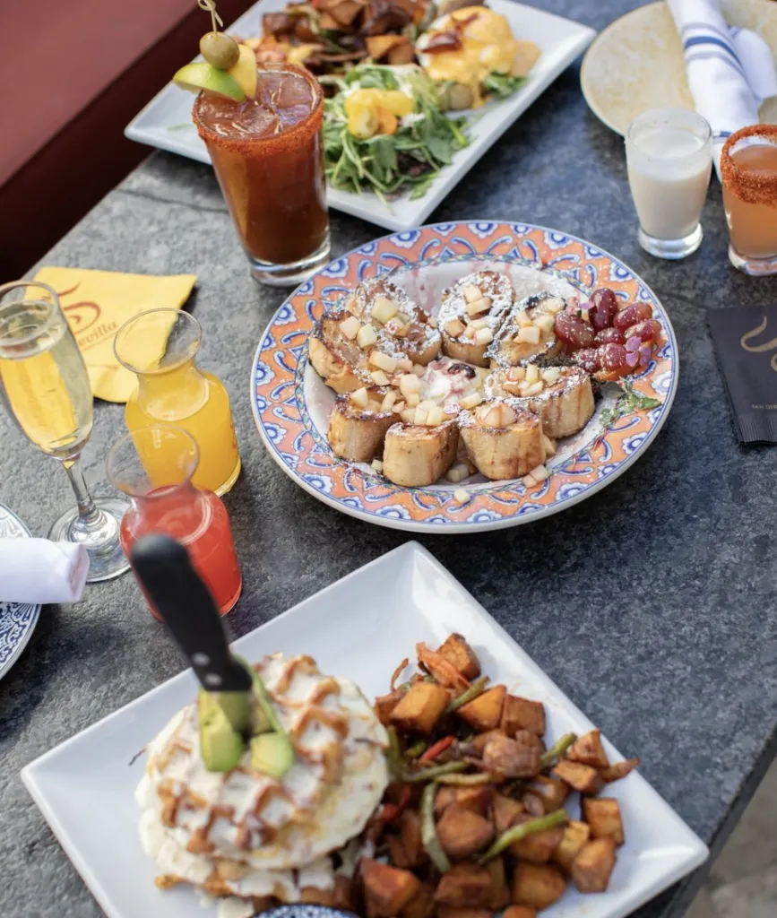 A table set with brunch foods including French toast with fruit, eggs benedict, breakfast potatoes, and a waffle topped with avocado and a knife, alongside cocktails and juice.