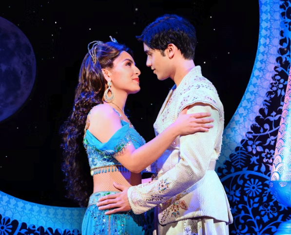 A woman in a turquoise, jeweled costume and a man in a white, ornate outfit gaze lovingly at each other on stage, under a moonlit night sky with intricate blue patterns in the background.