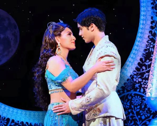 A woman in a turquoise, jeweled costume and a man in a white, ornate outfit gaze lovingly at each other on stage, under a moonlit night sky with intricate blue patterns in the background.