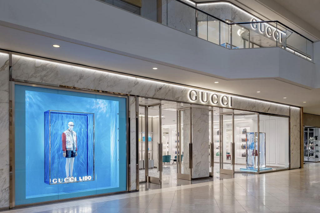 A modern Gucci store in a shopping mall with large glass windows, marble walls, and bright lighting. The storefront features a blue display with a mannequin dressed in stylish clothing.