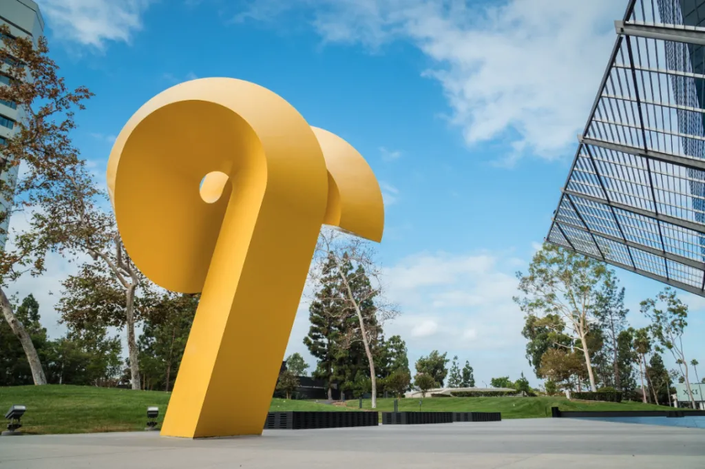 Large yellow abstract sculpture resembling a twisted ribbon stands outdoors on a paved area, surrounded by grass, trees, and modern buildings under a partly cloudy blue sky.