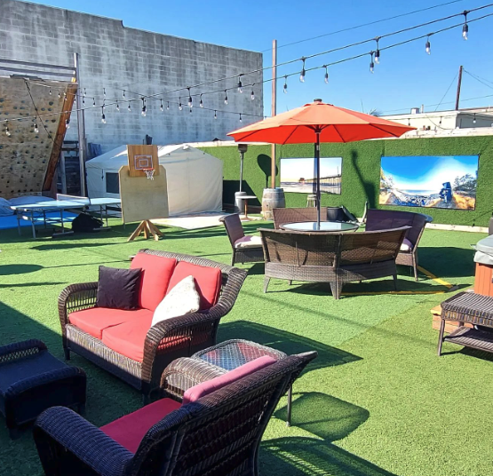 Outdoor patio with wicker furniture, a red cushioned sofa, chairs, a table with an orange umbrella, string lights, a basketball hoop, ping pong table, and wall art on artificial grass under a clear blue sky.