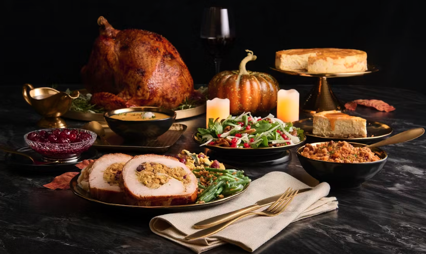 A Thanksgiving dinner spread with roast turkey, stuffed turkey slices, green beans, cranberries, stuffing, cornbread, a pumpkin centerpiece, candles, and a cheesecake on a dark table with gold utensils.