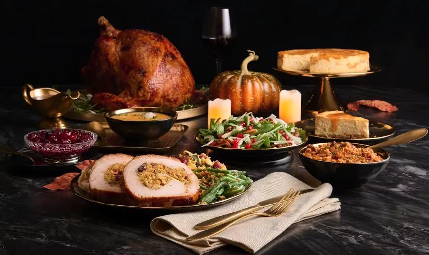 A Thanksgiving dinner spread with roast turkey, stuffed turkey slices, green beans, cranberries, stuffing, cornbread, a pumpkin centerpiece, candles, and a cheesecake on a dark table with gold utensils.