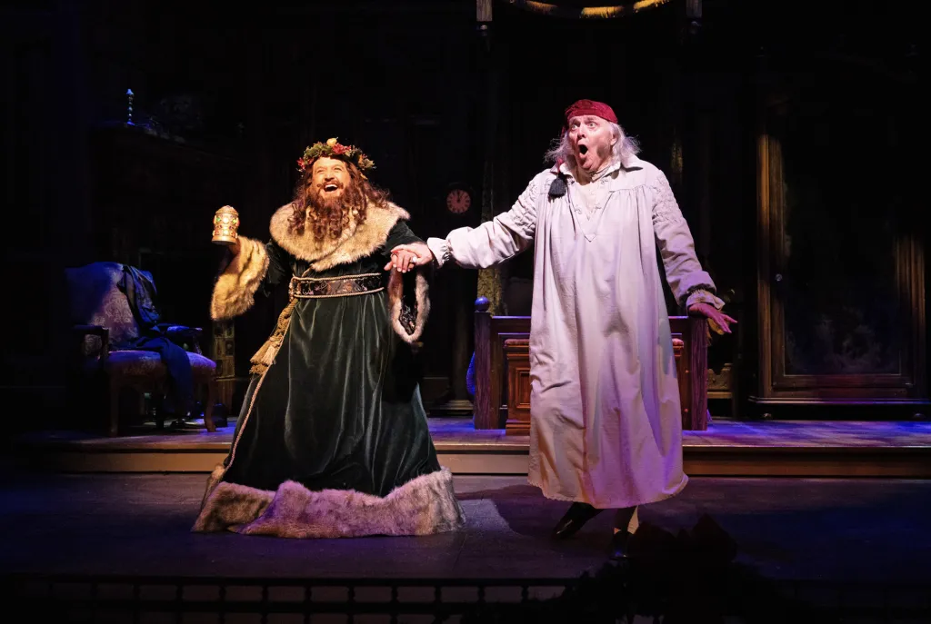 Two actors on stage: one in a festive, fur-trimmed green robe and wreath crown holding a goblet, the other in a white nightgown and cap, looking surprised. The set includes Victorian furniture and dark lighting.