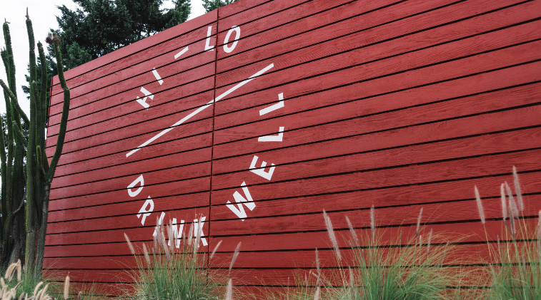 A large red wooden wall with the words DRINK WELL, HELLO written in white, arranged in a circular pattern. Desert plants and grasses grow at the base, with a tall cactus on the left.
