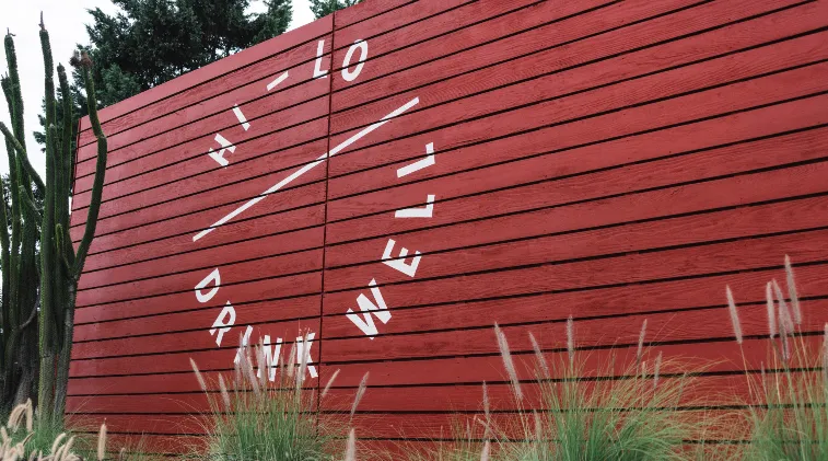 A large red wooden wall with the words DRINK WELL, HELLO written in white, arranged in a circular pattern. Desert plants and grasses grow at the base, with a tall cactus on the left.