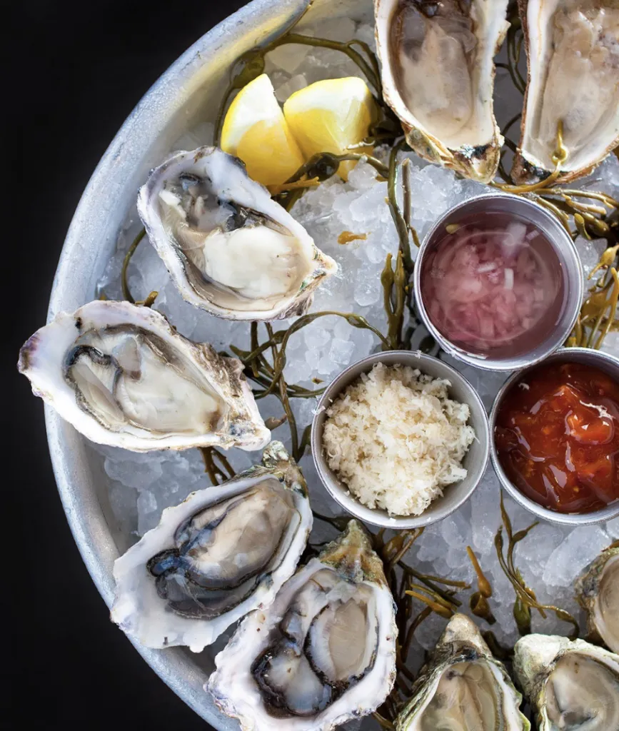 A round tray of raw oysters on ice, garnished with lemon wedges and seaweed, and served with metal cups of horseradish, cocktail sauce, and mignonette sauce.