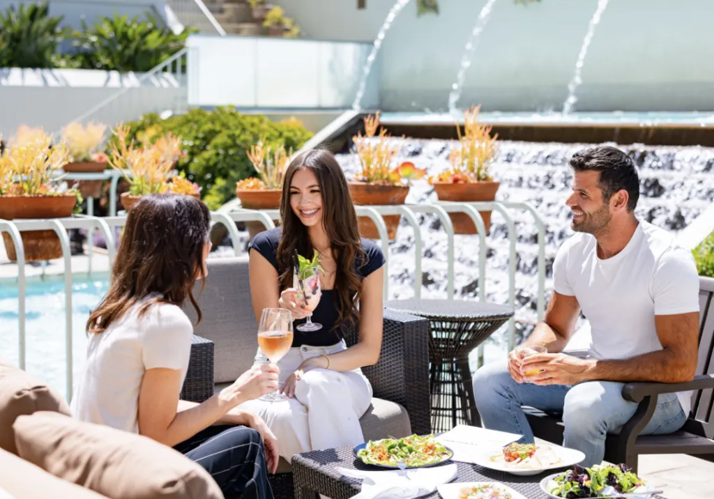 Three people sit outdoors by a fountain, enjoying drinks and food. Two women and one man are smiling and talking at a table with salads, pizza, and wine on a sunny day.