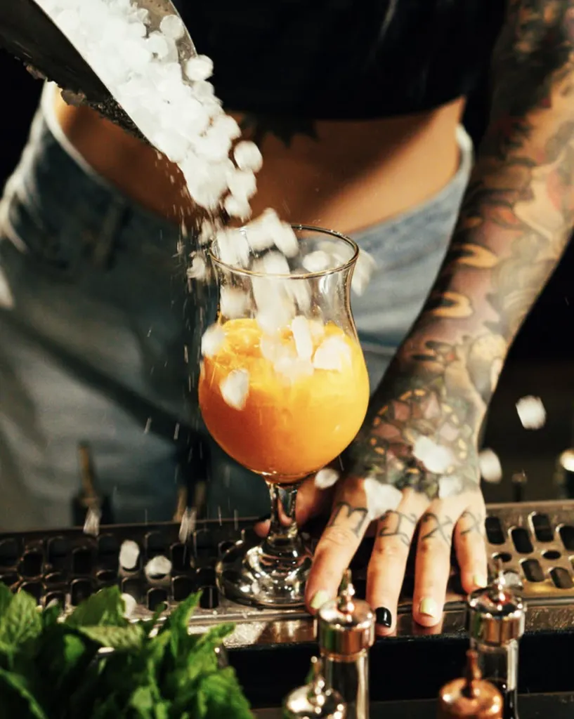 A tattooed bartender pours ice into a stemmed glass filled with an orange drink, with mint leaves and bar tools visible on the counter.