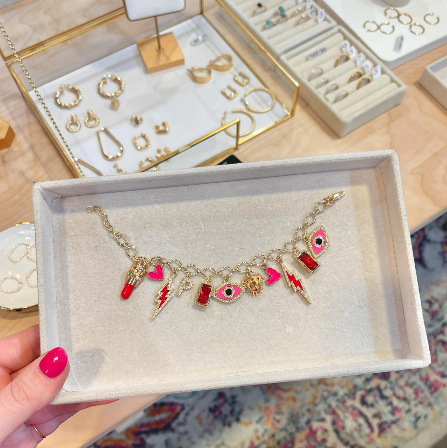 A hand with pink painted nails holds a jewelry box displaying a gold charm bracelet with colorful charms, including hearts, lips, a lightning bolt, and eye designs. Other jewelry pieces are visible on the table in the background.