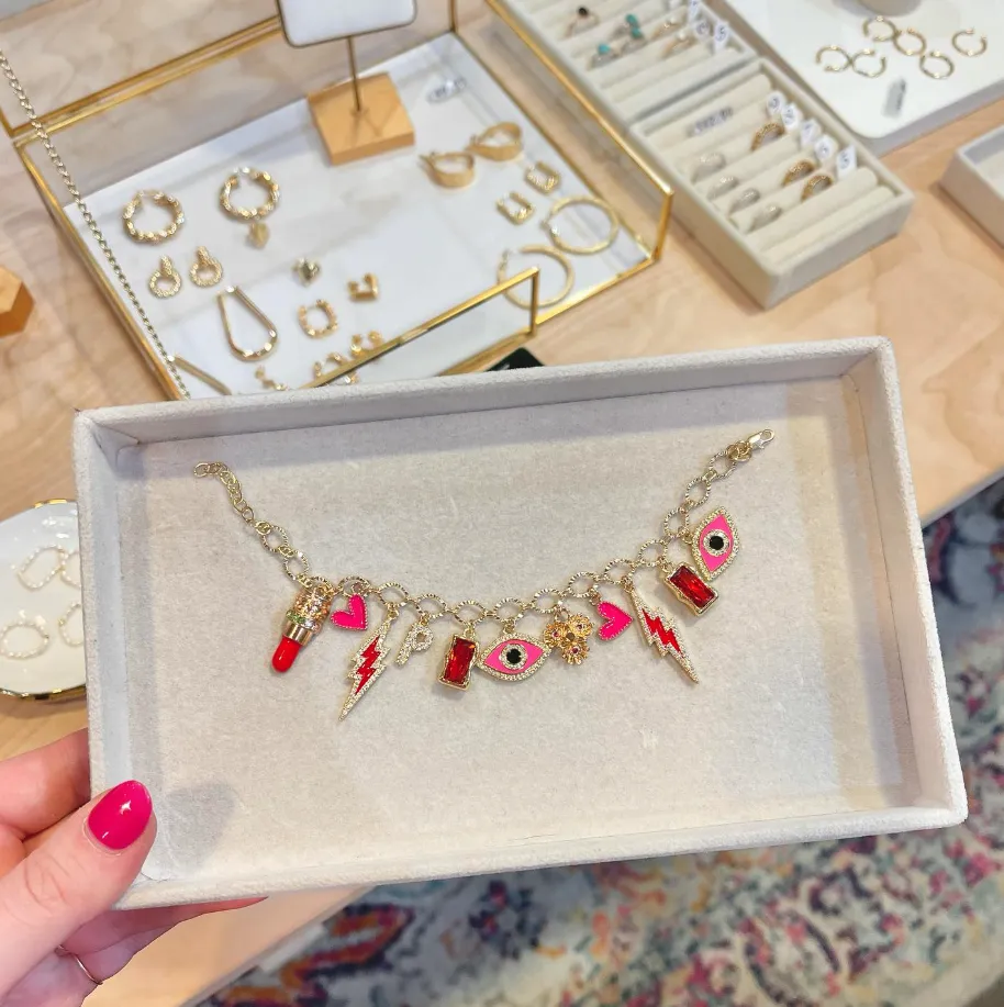 A hand with pink painted nails holds a jewelry box displaying a gold charm bracelet with colorful charms, including hearts, lips, a lightning bolt, and eye designs. Other jewelry pieces are visible on the table in the background.