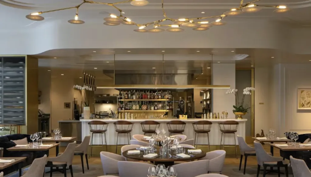 Chic, modern restaurant interior with round tables set for dining, elegant beige chairs, a stylish bar with high stools, and contemporary lighting hanging from the ceiling. Shelves with bottles and orchids decorate the bar area.