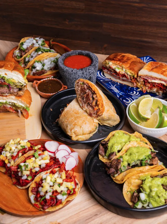 A colorful spread of Mexican food, including tacos with guacamole, tortas, burritos, sopes topped with diced veggies, lime wedges, and bowls of red and green salsa on a wooden table.