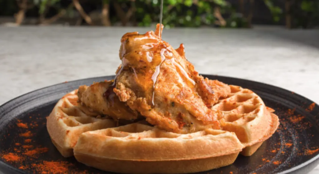 A piece of fried chicken sits atop a golden waffle on a black plate, while syrup is drizzled over the chicken. The dish is lightly dusted with red seasoning, and greenery is blurred in the background.