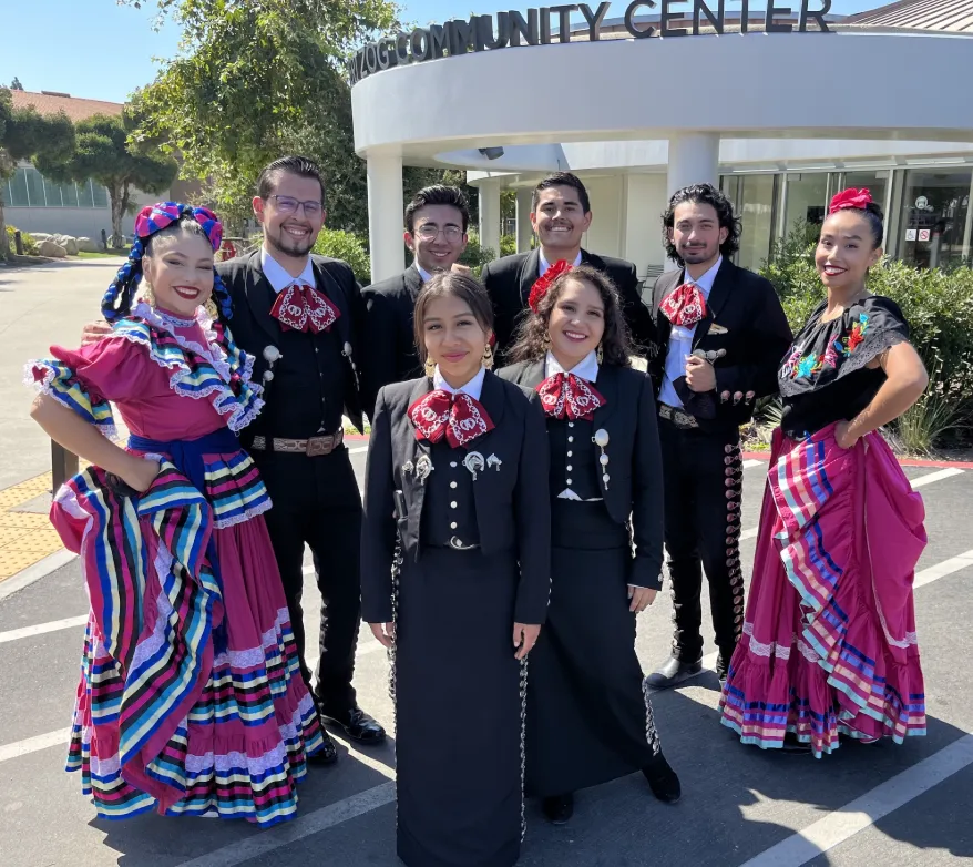 A group of eight people in colorful traditional mariachi attire pose and smile outside a community center. Four women wear vibrant skirts and blouses, while four men wear black suits with red bow ties.