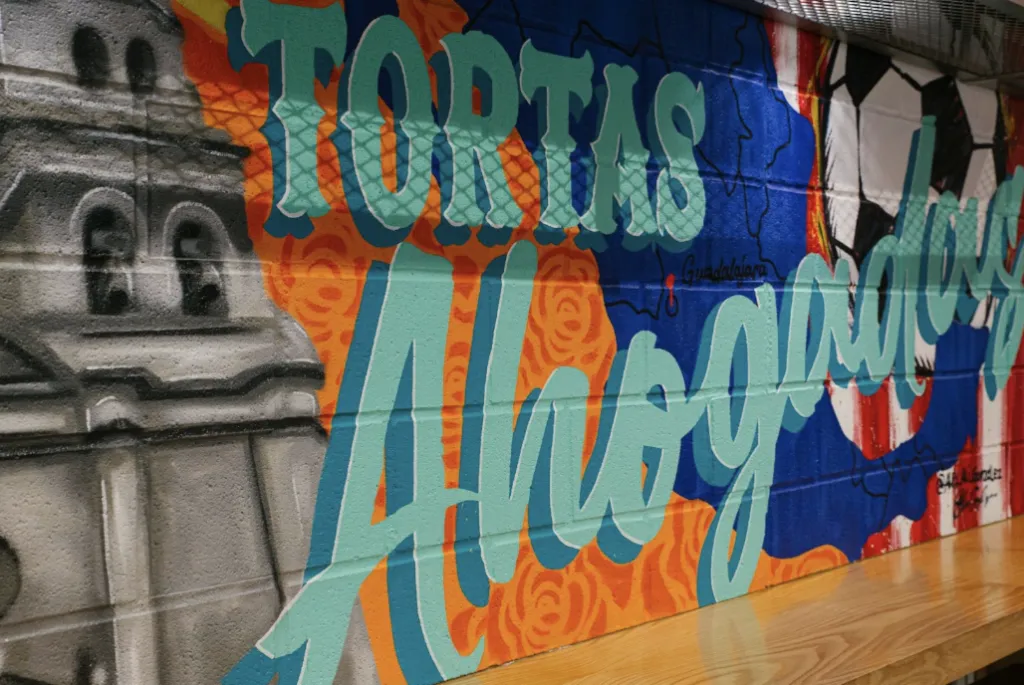 Colorful mural on a wall with large turquoise text reading “TORTAS Ahogadas,” surrounded by orange and blue patterns, a gray building on the left, and part of a soccer ball on the right.